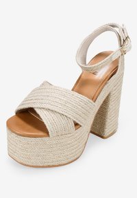 Beige rope wedge sandal featuring a cross-strap design, open toe, and adjustable ankle strap. Textured woven material with a wooden heel.