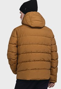 Braune Pufferjacke mit horizontalem Steppmuster, ausgestattet mit Kapuze und gerippten Akzenten. Getragen mit einer schwarzen Strickmütze.