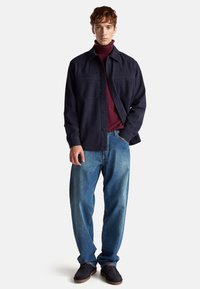 Granatowa koszula z guzikami, z dwiema kieszeniami na piersi, noszona na bordowym golfie. Jasnoniebieskie dżinsy w luźnym kroju, czarne buty z brązowymi podeszwami.