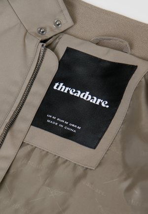 Béžová bunda na zip a se zapínáním na patentky, zobrazující černou štítek s logem "threadbare." a informacemi o velikosti: UK M, EUR M, USA M, vyrobeno v Číně.