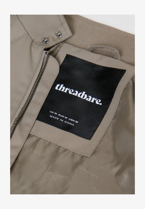 Béžová bunda na zip a se zapínáním na patentky, zobrazující černou štítek s logem "threadbare." a informacemi o velikosti: UK M, EUR M, USA M, vyrobeno v Číně.