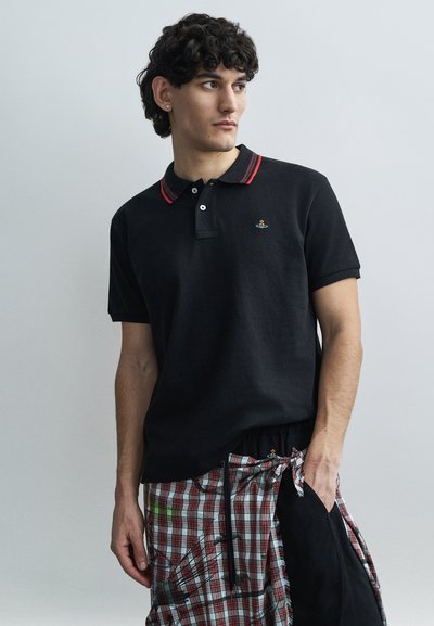 Young man with curly hair wearing a black polo shirt with red stripes on the collar and a plaid wrap skirt, looking to the side.