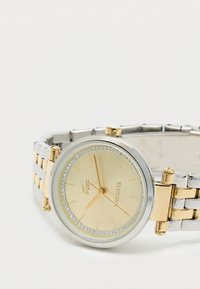 Kulaté, dvoubarevné hodinky se stříbrným nerezovým náramkem, zlatými akcenty, bezelkou posázenou krystaly a zlatým ciferníkem s tenkými ukazateli.