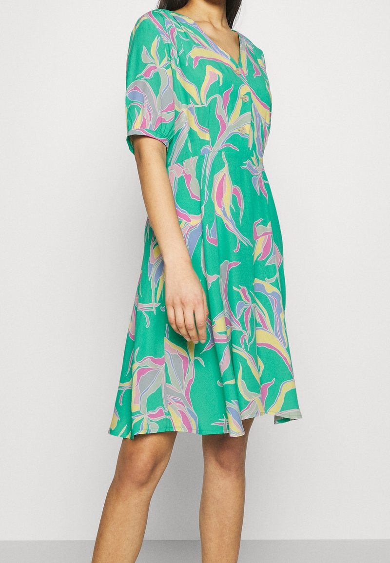 Femme portant une robe verte mi-longue avec un motif abstrait de feuilles rose, jaune et lavande et des manches courtes sur fond uni.
