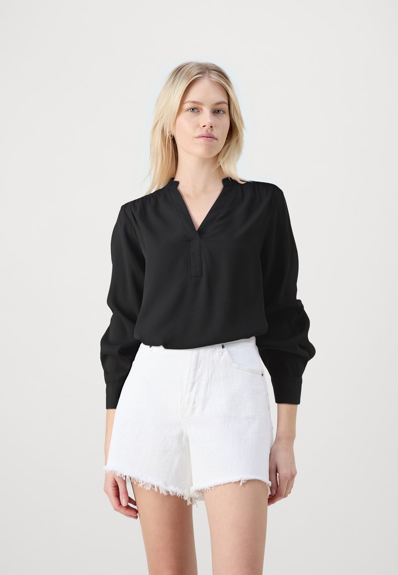 Schwarze Langarmbluse mit V-Ausschnitt, gerafften Schultern und lockerer Passform, kombiniert mit weißen, hochtaillierten Denim-Shorts mit fransigem Saum.
