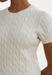 White short-sleeve cable-knit sweater featuring a ribbed crew neckline and subtle embroidered logo on the left chest.