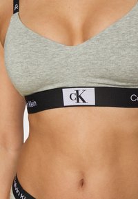 Heather grå myk bralette med bred sort elastisk bånd som viser "Calvin Klein"-logoen, med en sømløs design og V-hals.