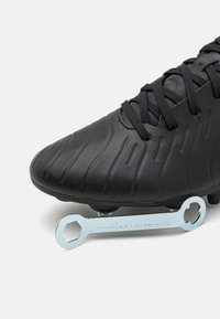 Czarne buty piłkarskie z teksturowaną powierzchnią i sznurówkami, umieszczone w towarzystwie metalowego klucza narzędziowego, który znajduje się pod korkami podeszwowymi.