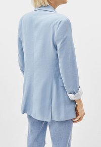 Světle modrý blazer se strukturovaným designem, měkkou texturou a dlouhými rukávy s bílými manžetami. Obsahuje zadní rozparek a boční kapsy.