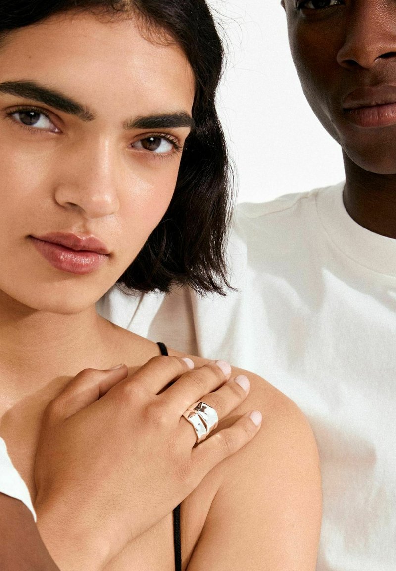 Hõbedast avaldus sõrmus lainelise disainiga, kantud käel, mis toetub paljale õlale. Nahatoonid on loomulikud; taust on valge ja lihtne.