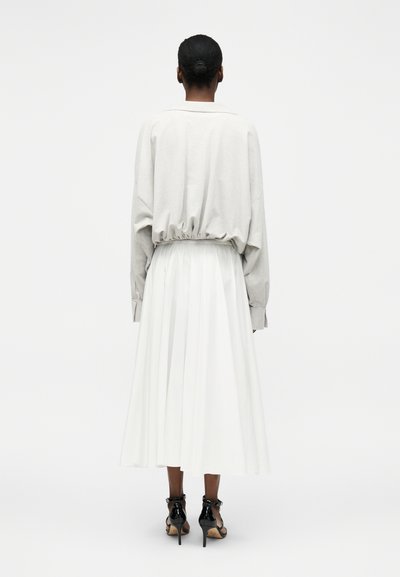 Γυναίκα με μαλλιά σε κότσο, φοράει ανοιχτό γκρι μακρυμάνικο τοπ, λευκή πλισέ midi φούστα και μαύρα ψηλοτάκουνα σανδάλια, στέκεται γυρισμένη με την πλάτη.
