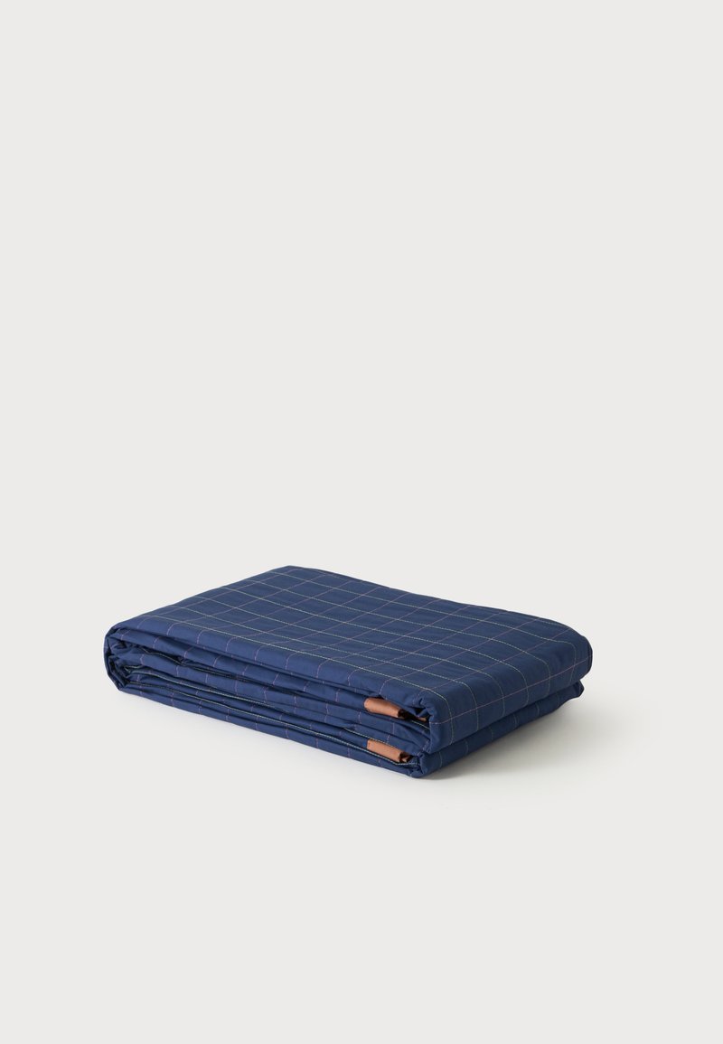 Couverture bleu foncé pliée avec un motif quadrillé fin et clair et de petites étiquettes en cuir sur fond blanc.