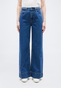 Γυναίκα φοράει ψηλόμεσα φαρδιά μπλε τζιν με πλεξούδα ζώνη και λευκά αθλητικά, στέκεται μπροστά σε απλό φωτεινό φόντο.