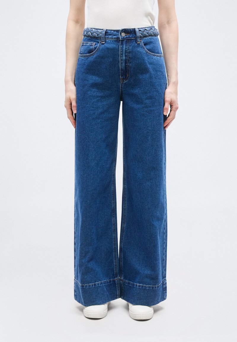 Γυναίκα φοράει ψηλόμεσα φαρδιά μπλε τζιν με πλεξούδα ζώνη και λευκά αθλητικά, στέκεται μπροστά σε απλό φωτεινό φόντο.