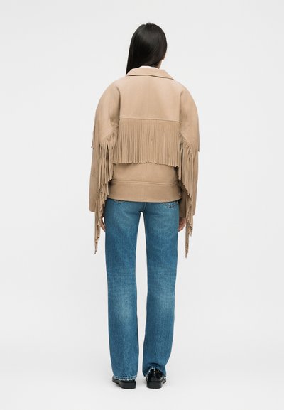 Vrouw die een beige suède jack draagt met franjes op de achterkant en mouwen, gecombineerd met een blauwe spijkerbroek en zwarte schoenen, staand met de rug naar de kijker.