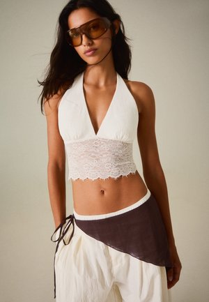 Kvinde iført store tonede solbriller, hvid blonder halterneck crop top og cremefarvede bukser med et mørkt asymmetrisk omslag bundet ved hoften.