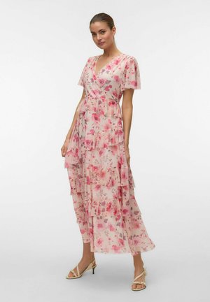 Kvinde iført en pink blomsterindpakket kjole med lagdelte flæser og korte ærmer, kombineret med beige sandaler med hæl og stropper, stående mod en hvid baggrund.