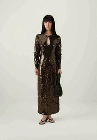 Vestido castanho de lantejoulas com mangas longas, decote em forma de buraco de fechadura, até ao chão, combinado com uma bolsa preta de palha e sandálias pretas.