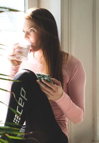 Woman sitting by a window, wearing a light pink long-sleeve top and black leggings with "cep" logo, holding a glass of water and a phone.