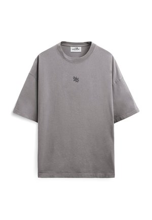 T-shirt gris surdimensionné à manches courtes avec un petit logo graphique noir centré sur la poitrine et une étiquette blanche au niveau du col.