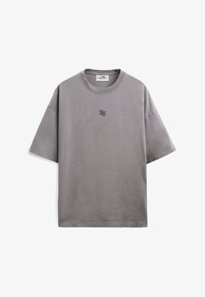 T-shirt grigia oversize a maniche corte con scollo rotondo, con piccolo logo grafico nero centrato sul petto e una etichetta bianca al collo.