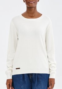 Vrouw in een eenvoudige witte lange mouwen trui en losse blauwe spijkerbroek tegen een effen witte achtergrond.