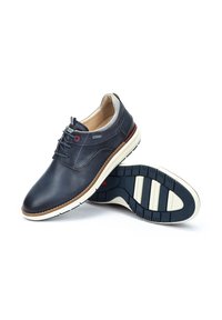Zapato de cuero azul marino con punta redonda, con acentos grises, diseño de cordones y suela texturizada en blanco y azul marino. Detalles rojos notables en los ojetes.