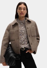 Chaqueta de lana marrón con dos bolsillos frontales, camisa estampada, vaqueros negros y un bolso negro texturizado. Clarice con cierres de botones y un collar.
