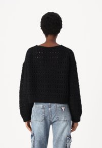 Svart, oversized strikket genser med åpen hekledesign, lange ermer, ribbet kant, brukt sammen med lyseblå cargojeans.