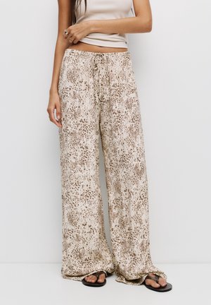 Vrouw met een beige mouwloze top, beige wijde broek met bruin luipaardprintje en zwarte sandalen tegen een effen witte achtergrond.