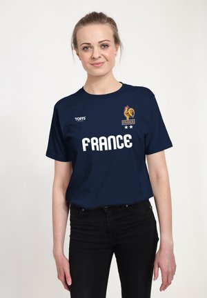 Frau mit hellem Haar, die ein marineblaues Frankreich-Sport-T-Shirt und schwarze Hose trägt und vor einem schlichten hellen Hintergrund steht.