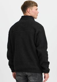 Veste en polaire noire avec une surface texturée, un col haut et une coupe décontractée. Présente un détail de couture subtil dans le dos.