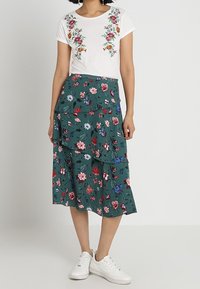 Vit t-shirt med blommig broderi och en grön, lager-på-lager blommig kjol med röda, blå och vita blommor. Enkla vita sneakers.