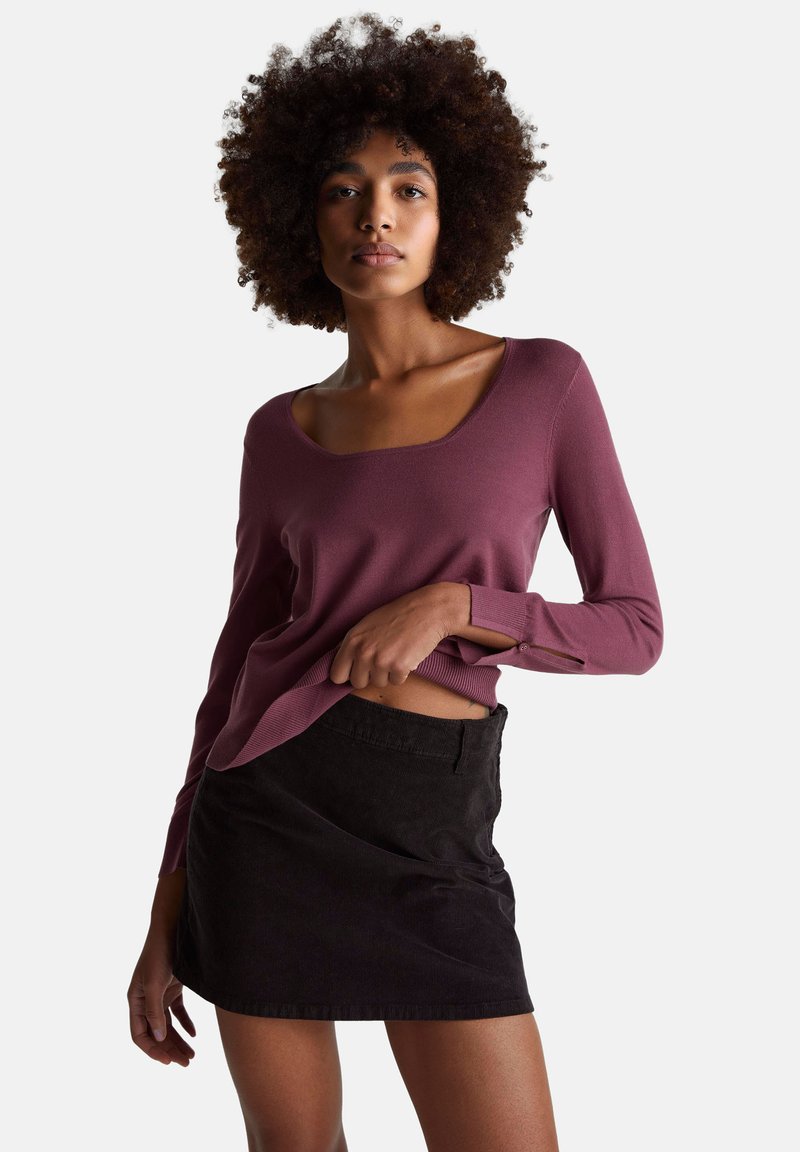 Frau mit lockigem Haar, die ein langärmliges Oberteil in Pflaumenfarbe und einen schwarzen Cord-Minirock trägt und das Oberteil leicht anhebt, um ihre Taille zu zeigen.