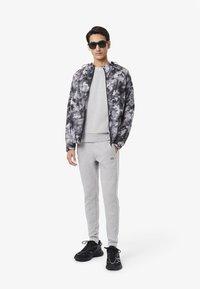 Un sweat-shirt gris et un pantalon de survêtement associés à une veste à motifs noirs et blancs. Des lunettes de soleil noires et des chaussures de sport noires complètent le look.