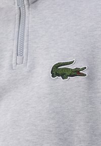 Pelēks zip-up džemperis ar teksturētu, izšūtu zaļu krokodila logotipu kreisajā krūtīs. Audums izskatās mīksts.