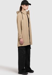 Chaqueta impermeable beige con capucha, cierre de cremallera y detalles de cordón, combinada con pantalones cargo negros y una gorra negra.