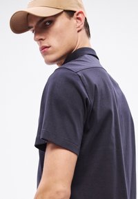 Tumesinine lühikeste varrukatega polosärk, millel on kontrastne krae. Kandmisega helepruun nokamüts. Sile kangas, minimaalne viimistlus, sportlik lõige.