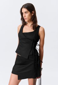 Haut court noir avec un décolleté carré, un ourlet asymétrique et des liens sur les côtés. Le tissu a une texture lisse avec un léger éclat. Associé à une mini jupe noire.