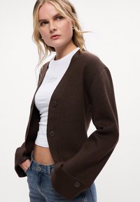 Hnedý cardigan s dizajnom do V, dlhými zvonovými rukávmi a dvoma tmavými gombíkmi. Nosený cez biele tričko s trblietkovým textom.