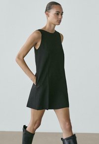 Czarna sukienka bez rękawów o prostej sylwetce, z bocznymi kieszeniami i gładką fakturą, dopełniona czarnymi kozakami za kolano.
