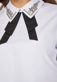 Weiße Bluse mit spitzem Kragen, verziert mit klaren Strasssteinen, und einem schwarzen Banddetail, das um den Hals gebunden ist. Glatte Stoffstruktur.