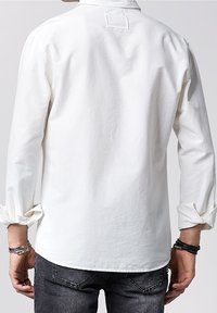 Camicia bianca in cotone con maniche lunghe, polsini con bottoni e dettaglio di cucitura sul retro. Presenta una vestibilità rilassata e una texture morbida.