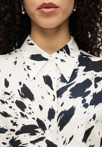 Λευκό πουκάμισο με αφηρημένο μοτίβο από μαύρες πιτσιλιές, μυτερός γιακάς και κούμπωμα με κουμπιά. Λείο ύφασμα με ελαφρά γυαλάδα.