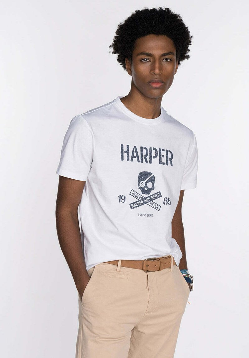 Camiseta blanca de algodón con un estampado gráfico azul que presenta una calavera y el texto "HARPER", combinada con pantalones beige y un cinturón marrón.