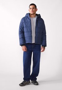 Chaqueta acolchada azul marino con capucha, cierre de cremallera y costuras horizontales, combinada con una camiseta gris y jeans azules, con zapatillas negras.