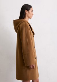 Manteau en laine marron avec une capuche, présentant une silhouette droite, de grands boutons à l'avant et des poches latérales. Texture lisse et design décontracté.