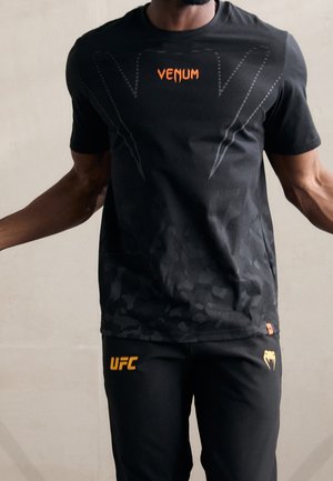 Mężczyzna ubrany w czarną odzież sportową Venum i UFC z pomarańczowymi logo, stojący z lekko uniesionymi ramionami na jednolitym tle.