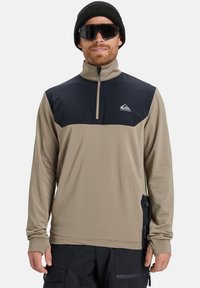 Beige langærmet pullover med en marineblå �øverste sektion, halv lynlås, struktureret stof og tommelfingerhuller ved ærmerne, parret med en sort beanie og solbriller.