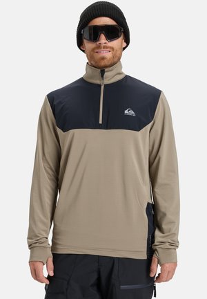 Pull en beige à manches longues avec une partie supérieure bleu marine, demi-zip, tissu texturé et trous pour les pouces aux poignets, associé à un bonnet noir et des lunettes de soleil.